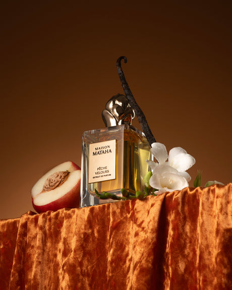 A bottle of Pêche Velours Extrait de Parfum by Fragrapedia Haus rests on orange velvet with a halved peach, white flower, and vanilla bean, capturing the essence of a velvety peach summer scent against a warm brown backdrop.