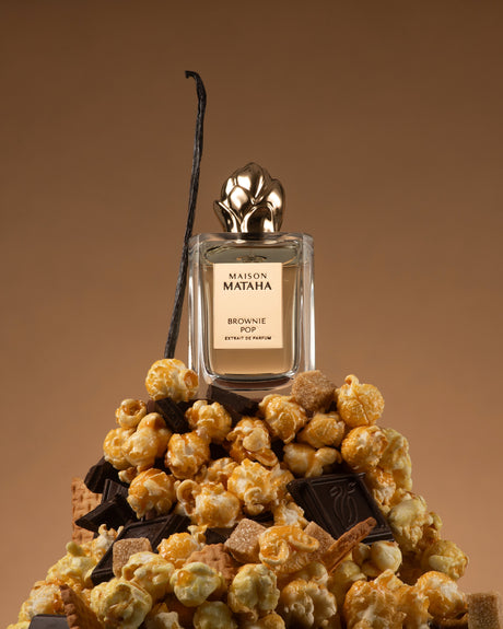 A bottle of Brownie Pop Extrait de Parfum by Maison Mataha rests atop caramel popcorn, chocolate pieces, wafer biscuits, and a vanilla bean, set against a rich brown background.
