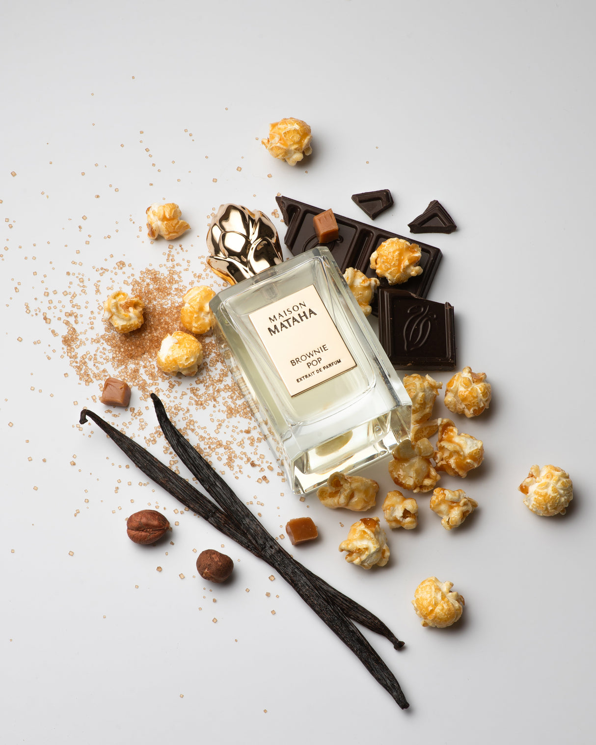 A bottle of Brownie Pop Extrait de Parfum by Maison Mataha sits on a white backdrop, surrounded by caramel popcorn, chocolate pieces, vanilla beans, and brown sugar—perfect for brownie fragrance enthusiasts.