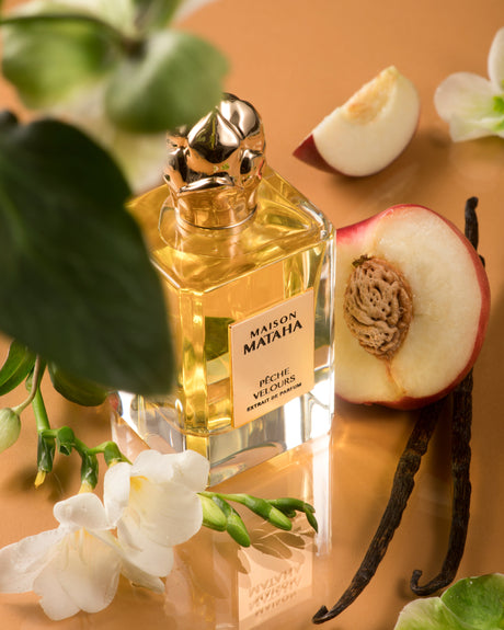 A bottle of Fragrapedia Haus Pêche Velours Extrait de Parfum by Maison Mataha stands on a peach surface among peach slices, vanilla beans, white flowers, and green leaves, suggesting a fresh and luxurious summer scent.