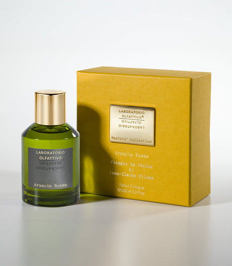 A green glass bottle with a gold cap labeled Arancia Rossa Eau de Parfum by Laboratorio Olfattivo stands beside a yellow box, both evoking Italian travels with fresh mandarin and bergamot notes.