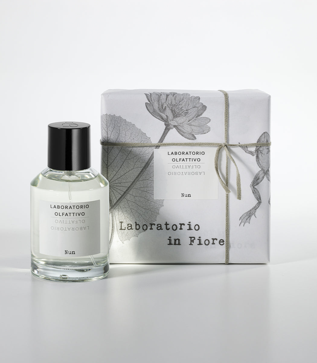 A glass bottle of Laboratorio Olfattivo's NUN Eau de Parfum stands beside a wrapped box with botanical designs, string, and matching branding, all set against a white background.