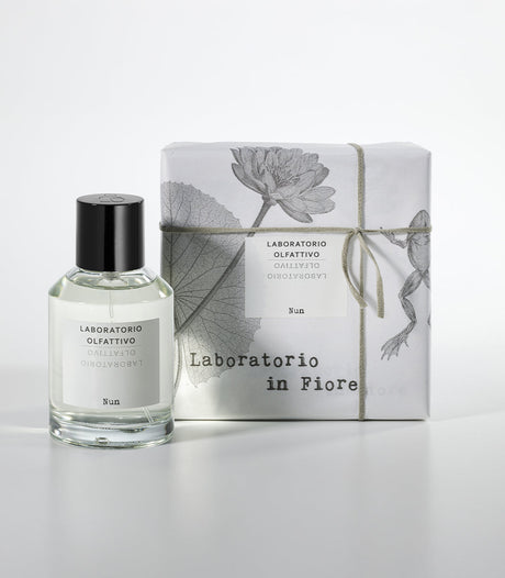 A glass bottle of Laboratorio Olfattivo's NUN Eau de Parfum stands beside a wrapped box with botanical designs, string, and matching branding, all set against a white background.