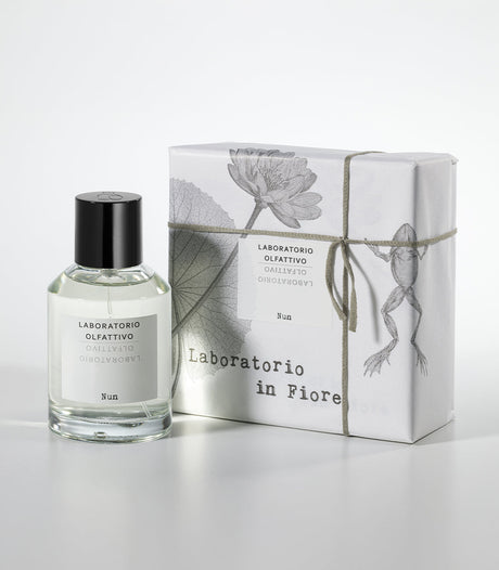 The NUN Eau de Parfum by Laboratorio Olfattivo is presented in a clear bottle beside its white box, adorned with floral and frog illustrations and tied with string—reflecting the lotus jasmine fragrance inside.
