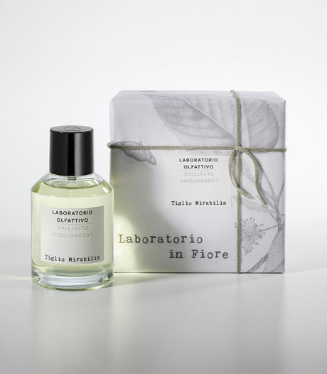A glass bottle of Tiglio Mirabilis Eau de Parfum by Laboratorio Olfattivo stands beside its white, botanically sketched box, evoking floral, spicy notes over a woody musky base in the Laboratorio in Fiore collection.