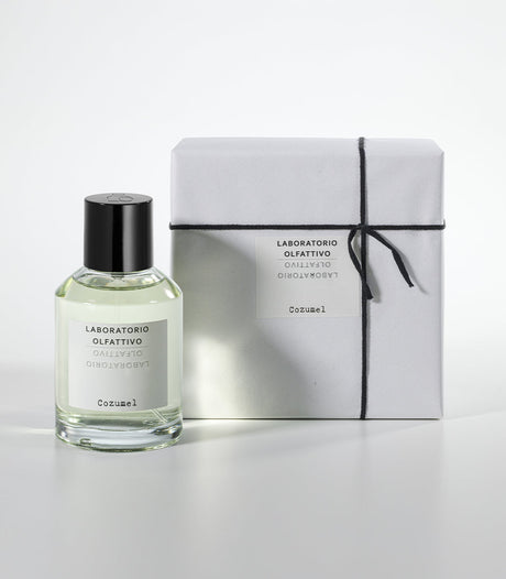 A clear glass bottle of Cozumel Eau de Parfum by Laboratorio Olfattivo, an aromatic men's fragrance, stands beside a white box tied with black string and featuring the matching Laboratorio Olfattivo label.