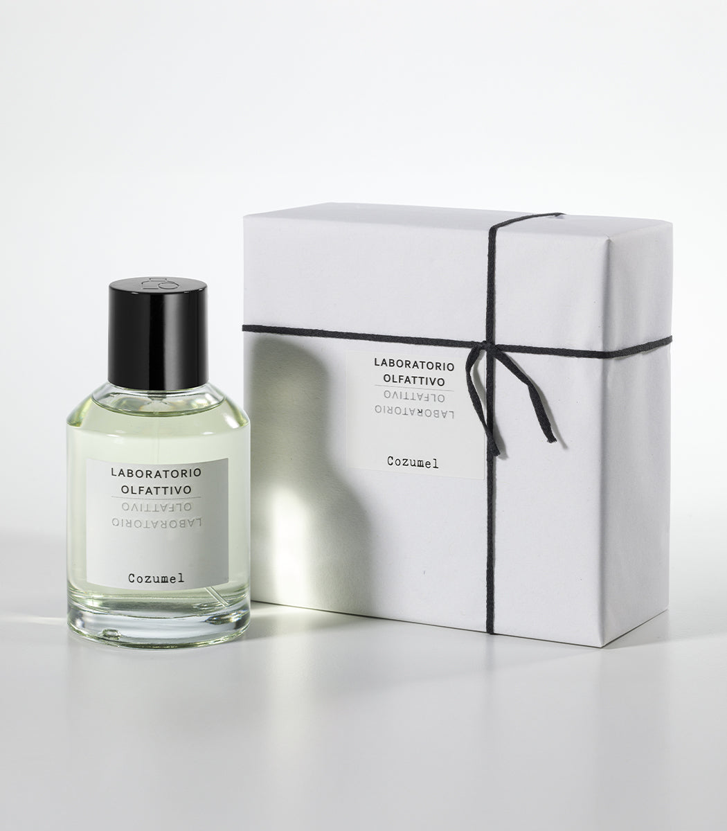 A clear bottle of Cozumel Eau de Parfum by Laboratorio Olfattivo stands beside a white gift box wrapped with black string and a matching label, both set against a plain white background.