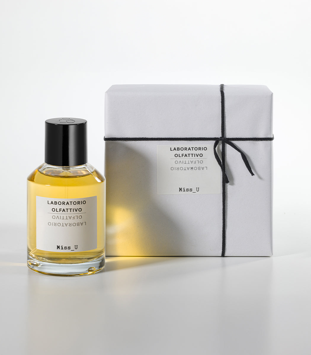 A bottle of Laboratorio Olfattivo MISS U Eau de Parfum with a black cap sits beside a gift-wrapped box tied with black string, both sporting minimalist white labels. Warm woods and ambery notes define the fragrance's allure.