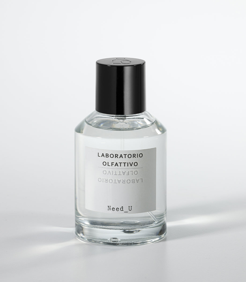 A clear glass bottle of NEED U Eau de Parfum by Laboratorio Olfattivo, with a black cap and labeled branding, rests on a white surface—an elegant unisex fragrance with soft jasmine notes.