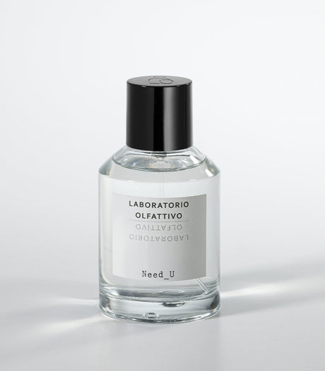A clear glass bottle of NEED U Eau de Parfum by Laboratorio Olfattivo, with a black cap and labeled branding, rests on a white surface—an elegant unisex fragrance with soft jasmine notes.