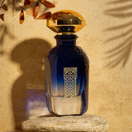 A luxurious blue Granada Extrait de Parfum bottle by Fragrapedia Haus with a gold cap stands on stone amid sunlight and brown leaves, evoking soft notes of rose and juniper for an elegant, inviting atmosphere.