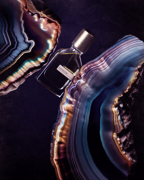 A perfume bottle with a gold cap rests on a dark surface, encircled by vivid agate slices in blue, purple, and orange. The label reads "Risvelium Eau de Parfum - Orto Parisi" by Fragrapedia Haus—an extraordinary, ethereal fragrance.