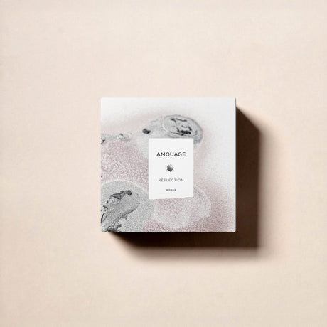 A square perfume box from Fragrapedia Haus, labeled Reflection Woman Eau de Parfum - Amouage, rests on a light beige surface. Its minimalist design features abstract gray and pale pink patterns, evoking the white floral scent inside.