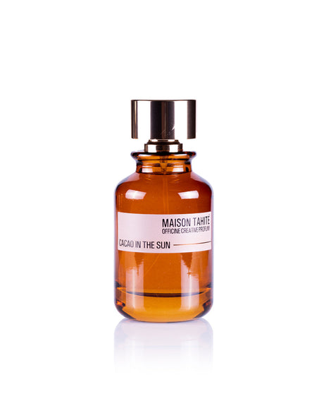 A brown glass bottle with a metallic cap, labeled "Cacao in the Sun Eau de Parfum" by Maison Tahite, sits on a white background, evoking a rich gourmand scent with distinctive cocoa notes.