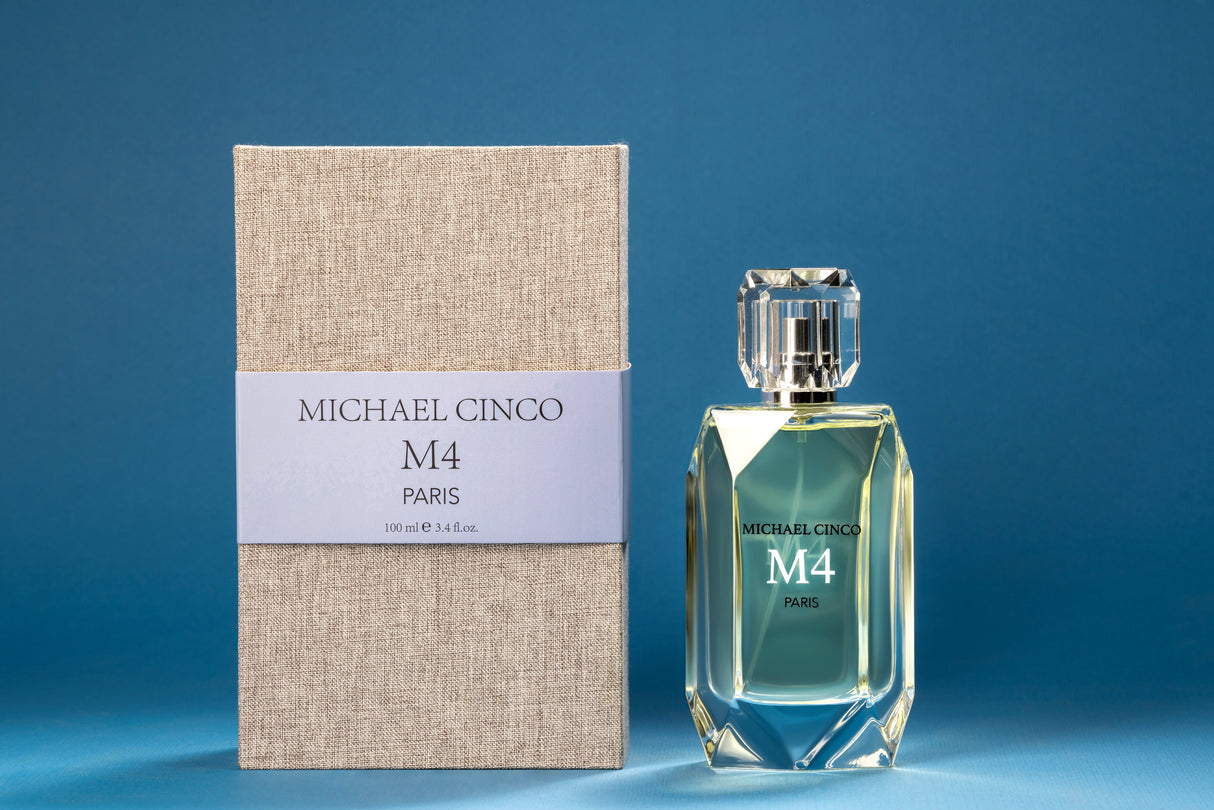 A faceted bottle of Fragrapedia Haus M4 Eau de Parfum by Michael Cinco, with marine notes, stands beside its textured beige box and blue label against a solid blue background, reflecting light elegantly.