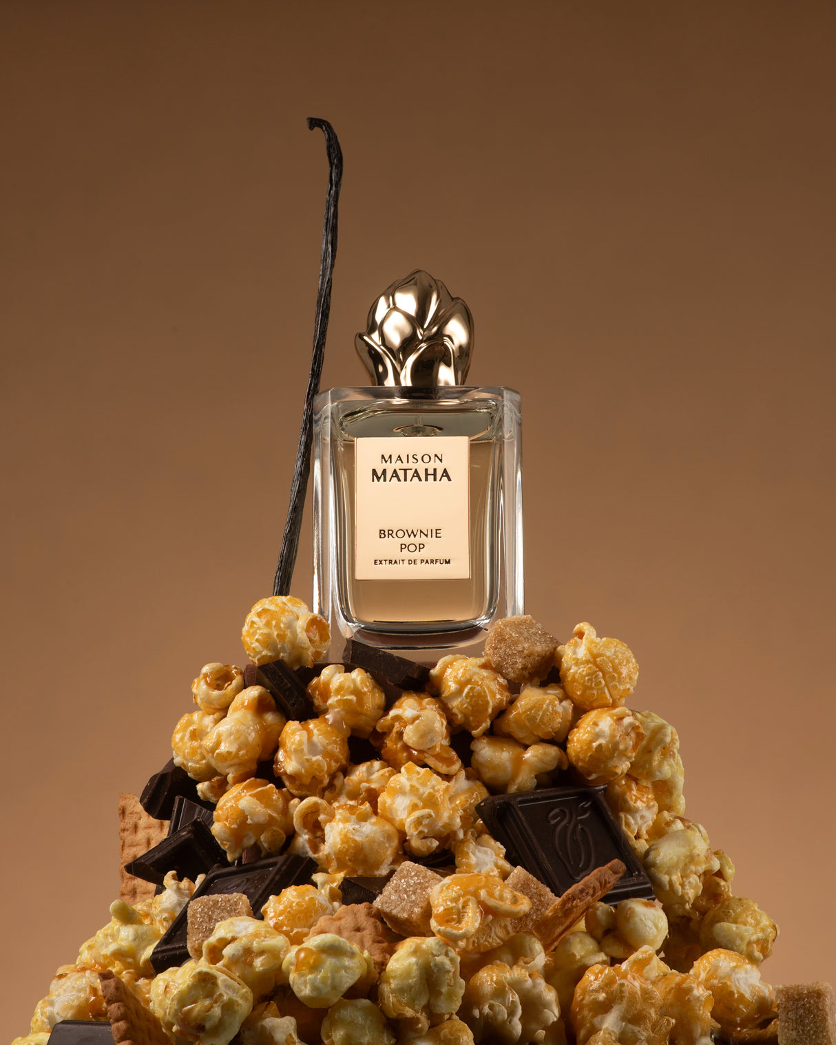A bottle of Brownie Pop Extrait de Parfum by Maison Mataha rests atop caramel popcorn, chocolate pieces, wafer biscuits, and a vanilla bean, set against a rich brown background.