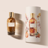 A glass bottle of Almond Eau de Parfum by Fragrapedia Haus, with a gold cap, stands beside its cylindrical packaging adorned with floral and almond designs on a light background, reflecting the creamy, rich oriental scent inside.