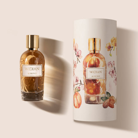 A glass bottle of Almond Eau de Parfum by Fragrapedia Haus, with a gold cap, stands beside its cylindrical packaging adorned with floral and almond designs on a light background, reflecting the creamy, rich oriental scent inside.