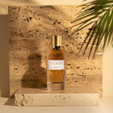 Fragrapedia Haus Almond Eau de Parfum by Widian, in a glass bottle with a gold cap, sits on a light stone platform against a textured stone backdrop and palm leaf shadow, reflecting the creamy richness of this elegant almond scent.