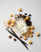 A bottle of Brownie Pop Extrait de Parfum by Maison Mataha sits on a white backdrop, surrounded by caramel popcorn, chocolate pieces, vanilla beans, and brown sugar—perfect for brownie fragrance enthusiasts.