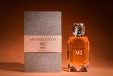 A bottle of Fragrapedia Haus M2 Eau de Parfum by Michael Cinco, filled with amber liquid, stands by its textured gray box on a brown surface, reflecting an exquisite fragrance inspired by oriental spirituality.