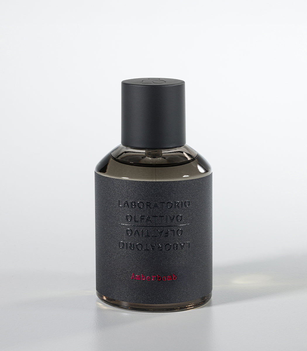 A dark, cylindrical Amberbomb Eau de Parfum bottle by Laboratorio Olfattivo features a matte black cap and textured gray label with silver brand text and red "Amberbomb," embodying the refined luxury of the Extreme Collection.