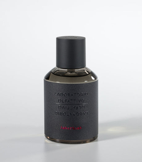 A dark, cylindrical Amberbomb Eau de Parfum bottle by Laboratorio Olfattivo features a matte black cap and textured gray label with silver brand text and red "Amberbomb," embodying the refined luxury of the Extreme Collection.
