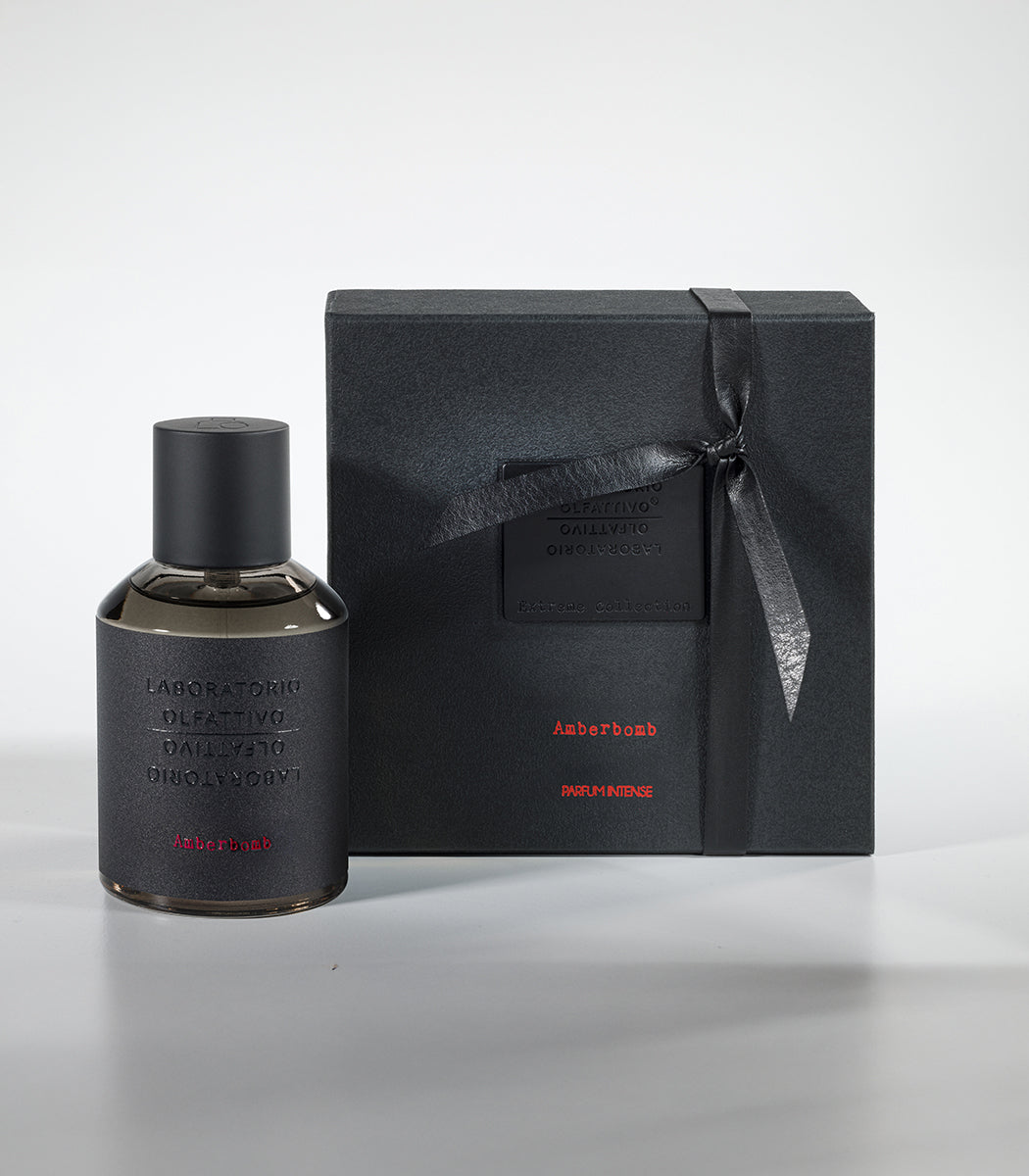 The Amberbomb Eau de Parfum by Laboratorio Olfattivo stands next to a sleek black gift box with red accents and subtle embossed text, both reflecting minimalist luxury on a light gray background.