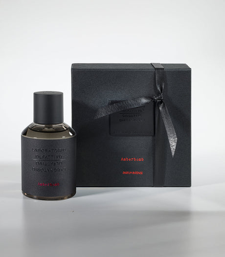 The Amberbomb Eau de Parfum by Laboratorio Olfattivo stands next to a sleek black gift box with red accents and subtle embossed text, both reflecting minimalist luxury on a light gray background.