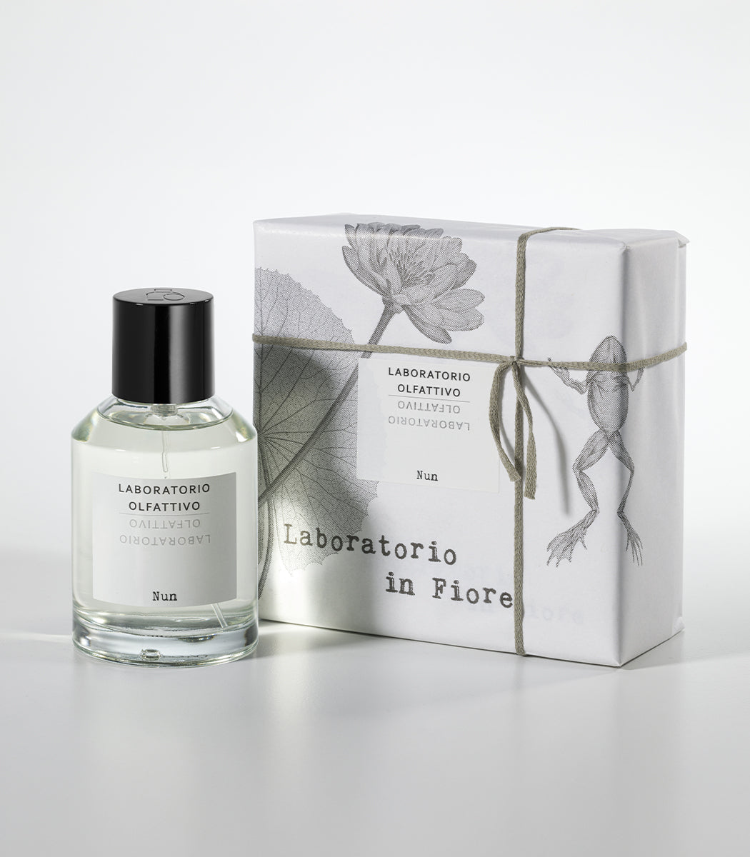 The NUN Eau de Parfum by Laboratorio Olfattivo is presented in a clear bottle beside its white box, adorned with floral and frog illustrations and tied with string—reflecting the lotus jasmine fragrance inside.
