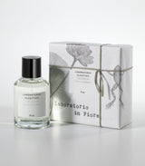 The NUN Eau de Parfum by Laboratorio Olfattivo is presented in a clear bottle beside its white box, adorned with floral and frog illustrations and tied with string—reflecting the lotus jasmine fragrance inside.
