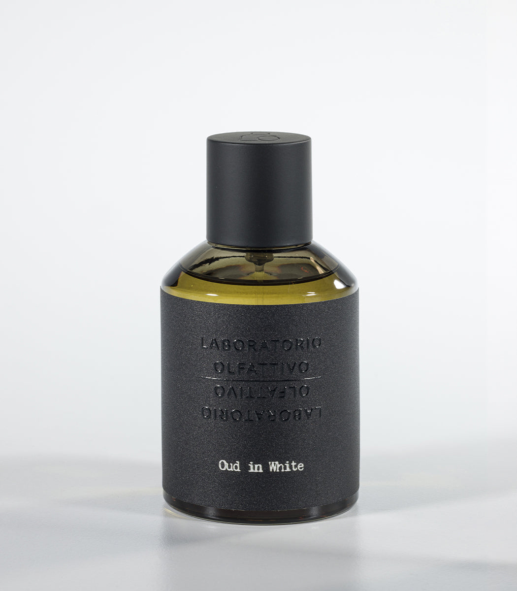 A bottle of Oud in White Eau de Parfum by Fragrapedia Haus, featuring a black cylindrical cap and textured dark gray label against a light background—an elegant floral fragrance with soft sandalwood notes.