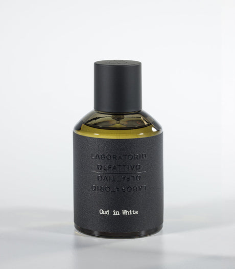 A bottle of Oud in White Eau de Parfum by Fragrapedia Haus, featuring a black cylindrical cap and textured dark gray label against a light background—an elegant floral fragrance with soft sandalwood notes.
