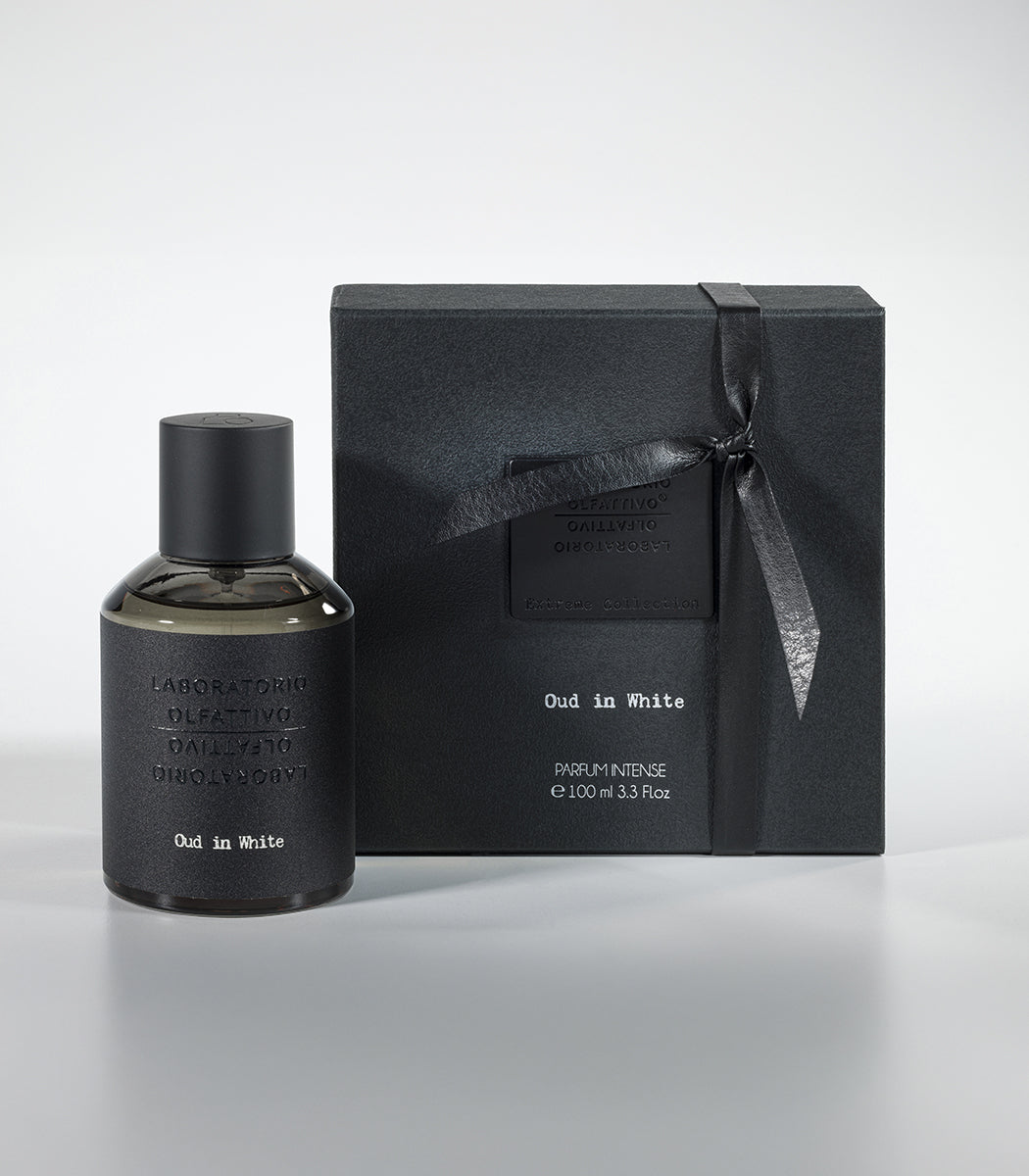 A bottle of Oud in White Eau de Parfum by Fragrapedia Haus sits beside its matching black ribbon-tied gift box, both arranged on a light gray surface.