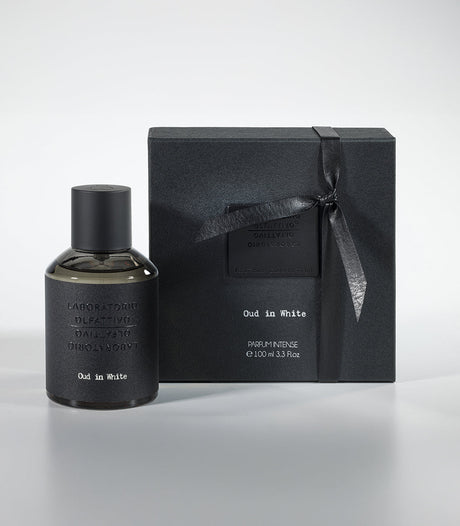 A bottle of Oud in White Eau de Parfum by Fragrapedia Haus sits beside its matching black ribbon-tied gift box, both arranged on a light gray surface.