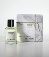 A glass bottle of Tiglio Mirabilis Eau de Parfum by Laboratorio Olfattivo stands beside its white, botanically sketched box, evoking floral, spicy notes over a woody musky base in the Laboratorio in Fiore collection.