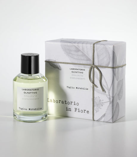 A bottle of Laboratorio Olfattivo's Tiglio Mirabilis Eau de Parfum stands beside its illustrated box, tied with string; the fragrance fuses spicy and floral notes atop a woody musky base.