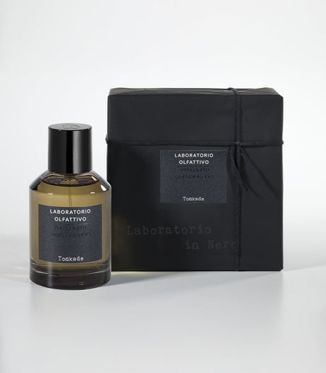 A bottle of Laboratorio Olfattivo Tonkade Eau de Parfum, featuring warm Tonka Bean notes, stands beside its matching black gift-wrapped box with a label, both set on a white surface against a neutral background.