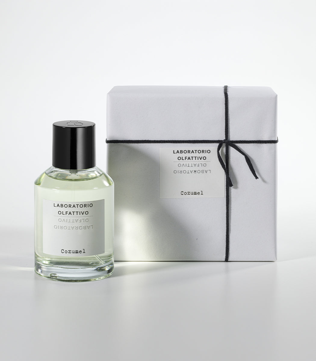 A clear glass bottle of Cozumel Eau de Parfum by Laboratorio Olfattivo, an aromatic men's fragrance, stands beside a white box tied with black string and featuring the matching Laboratorio Olfattivo label.