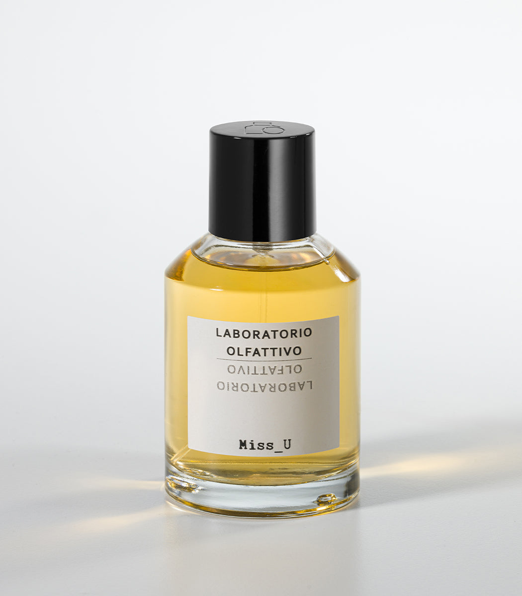 A clear glass bottle of MISS U Eau de Parfum by Laboratorio Olfattivo, filled with yellow liquid and topped with a black cap, stands against a white background—an amber fragrance capturing warm woods and sensual notes.