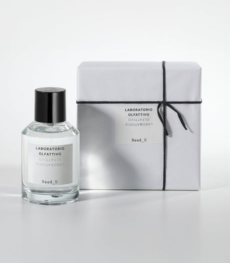 A clear glass bottle of NEED U Eau de Parfum by Laboratorio Olfattivo, a refined unisex scent featuring Ambroxan and jasmine notes, stands beside its white box tied with black string against a plain white background.