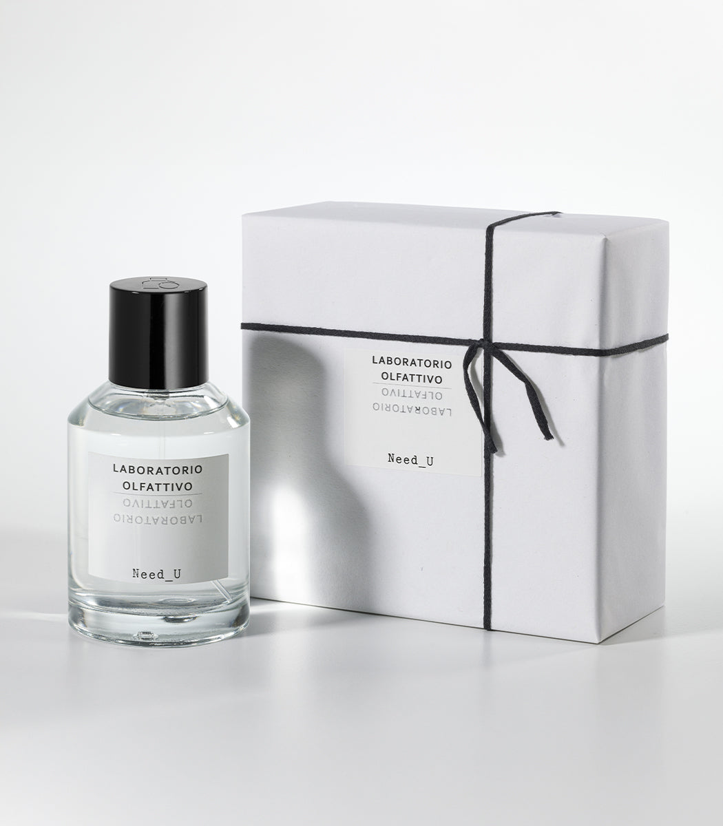 A bottle of Laboratorio Olfattivo NEED U Eau de Parfum stands beside a white, neatly wrapped box tied with black string on a light gray background.
