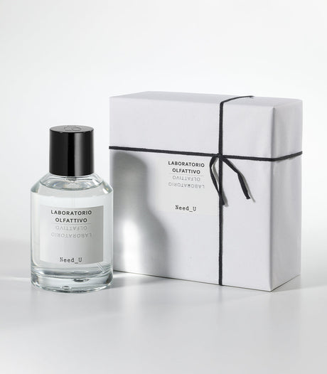 A bottle of Laboratorio Olfattivo NEED U Eau de Parfum stands beside a white, neatly wrapped box tied with black string on a light gray background.