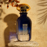 A luxurious blue Granada Extrait de Parfum bottle by Fragrapedia Haus with a gold cap stands on stone amid sunlight and brown leaves, evoking soft notes of rose and juniper for an elegant, inviting atmosphere.