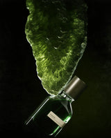A green bottle of Viridi Eau de Parfum by Fragrapedia Haus rests beneath a textured green crystal, evoking herbal Yemeni traditions, all dramatically lit against a dark backdrop.