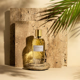 A bottle of Fragrapedia Haus White Eau de Parfum by Widian, gold-capped, rests on stone amid palm shadows and textured tiles, evoking warm light and reflecting a floral bouquet with hints of precious woods and musk.