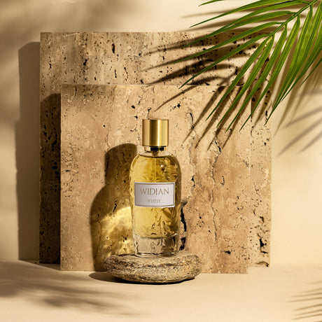 A bottle of Fragrapedia Haus White Eau de Parfum by Widian, gold-capped, rests on stone amid palm shadows and textured tiles, evoking warm light and reflecting a floral bouquet with hints of precious woods and musk.