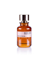 A brown glass bottle with a metallic cap, labeled "Cacao in the Sun Eau de Parfum" by Maison Tahite, sits on a white background, evoking a rich gourmand scent with distinctive cocoa notes.